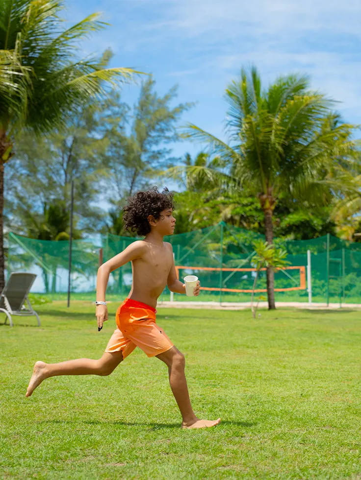 Área externa do resort com criança correndo na grama, ao fundo espreguiçadeiras, uma quadra de areia e muito coqueiros em dia ensolarado.