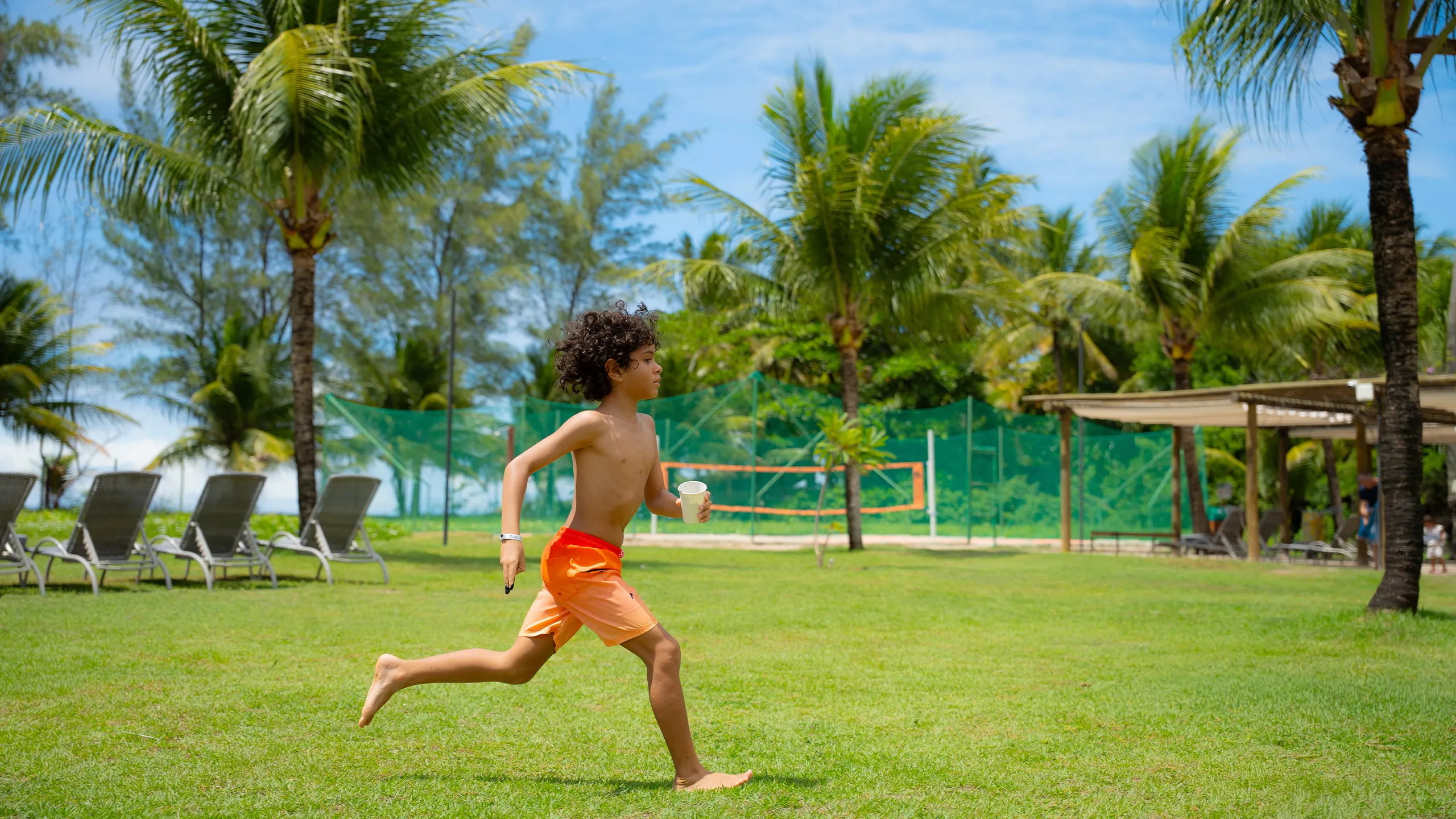 Área externa do resort com criança correndo na grama, ao fundo espreguiçadeiras, uma quadra de areia e muito coqueiros em dia ensolarado.