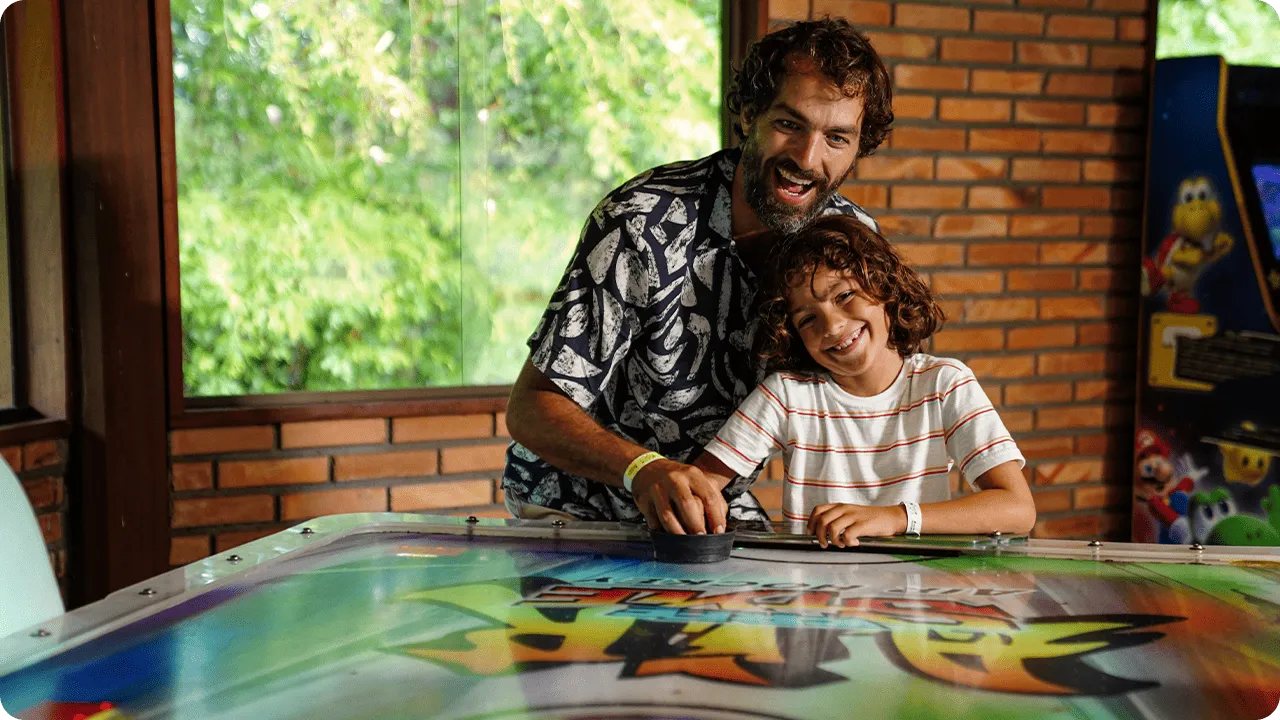 Pai e filho sorrindo enquanto jogam air hockey na área de jogos do resort.