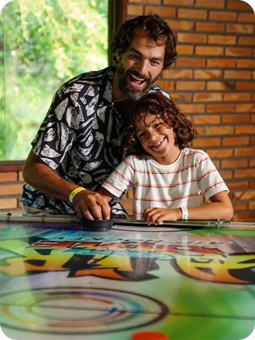 Pai e filho sorrindo enquanto jogam air hockey na área de jogos do resort.