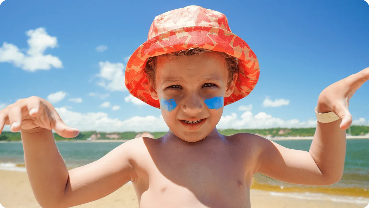 Criança sorridente com chapéu colorido e protetor solar no rosto na praia