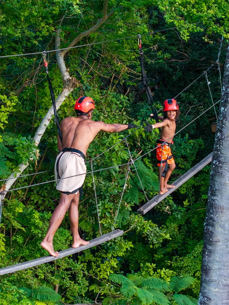 Pai e filho praticando arvorismo no resort. Em trilha suspensa entre as árvores altas, e bastante verde ao fundo.