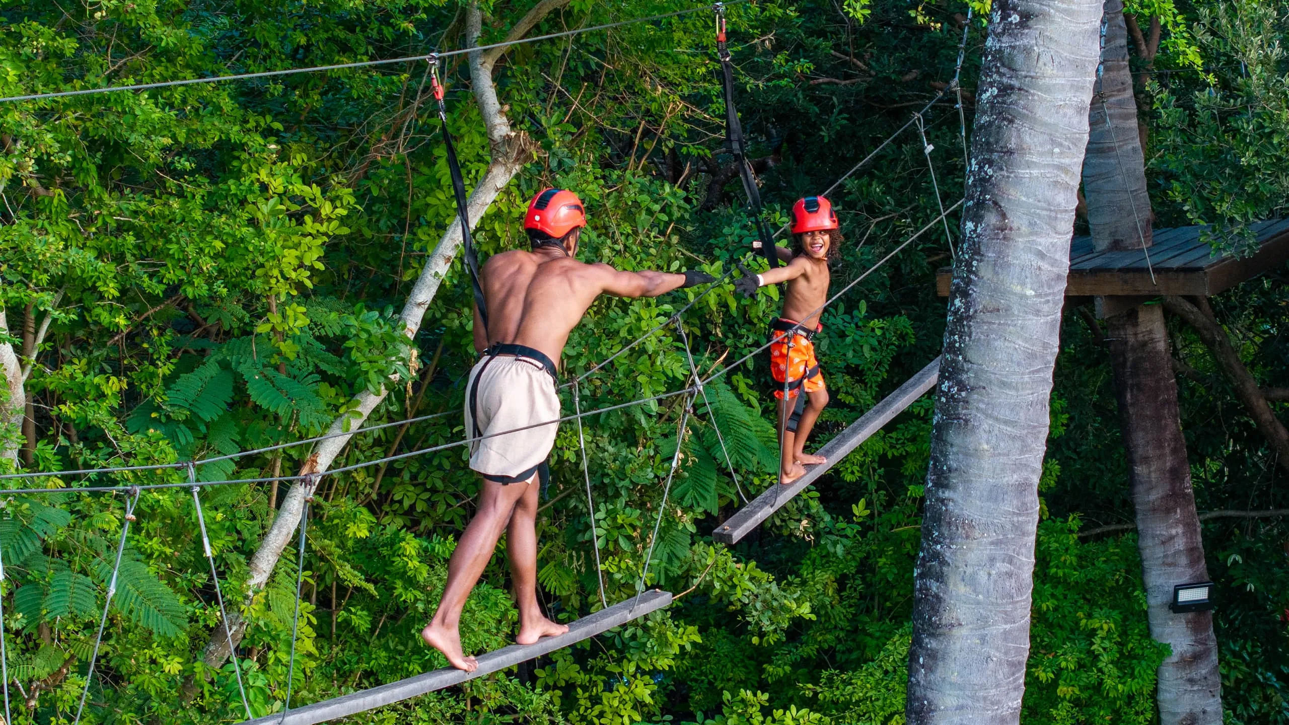 Pai e filho praticando arvorismo no resort. Em trilha suspensa entre as árvores altas, e bastante verde ao fundo.