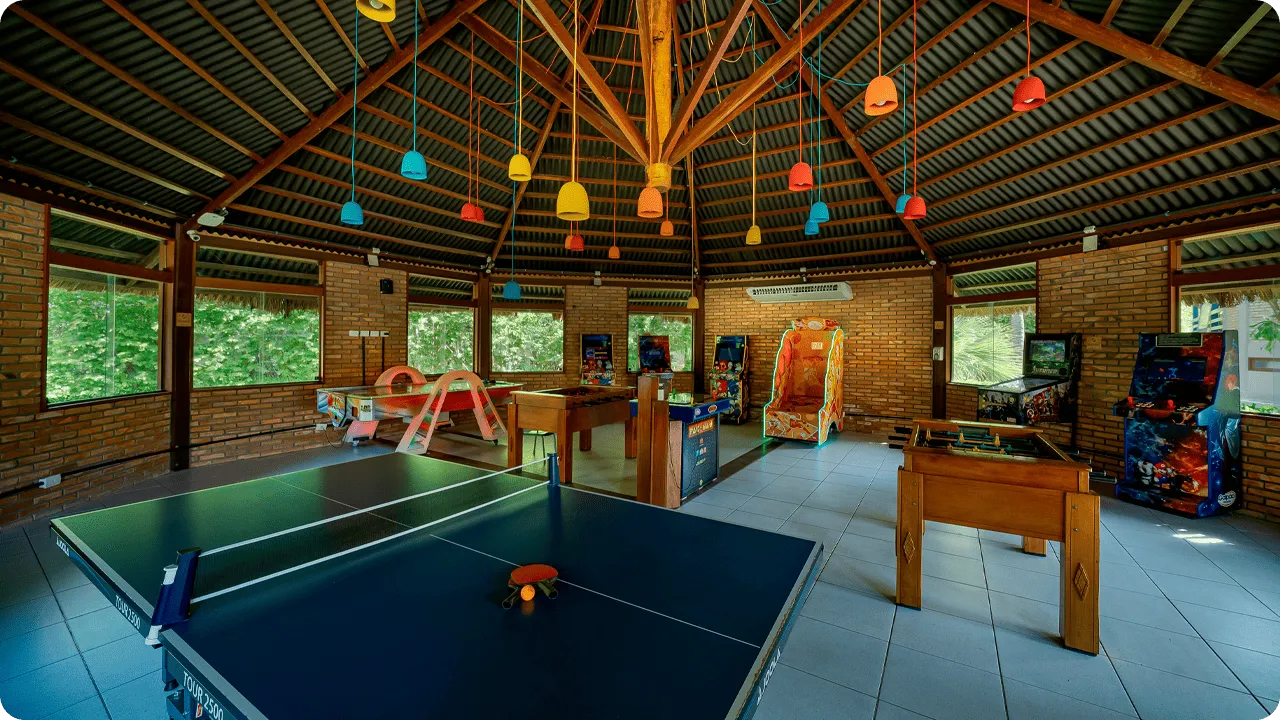Espaço de jogos com mesas de pingue-pongue, pebolim, fliperamas e brinquedos infantis em resort.