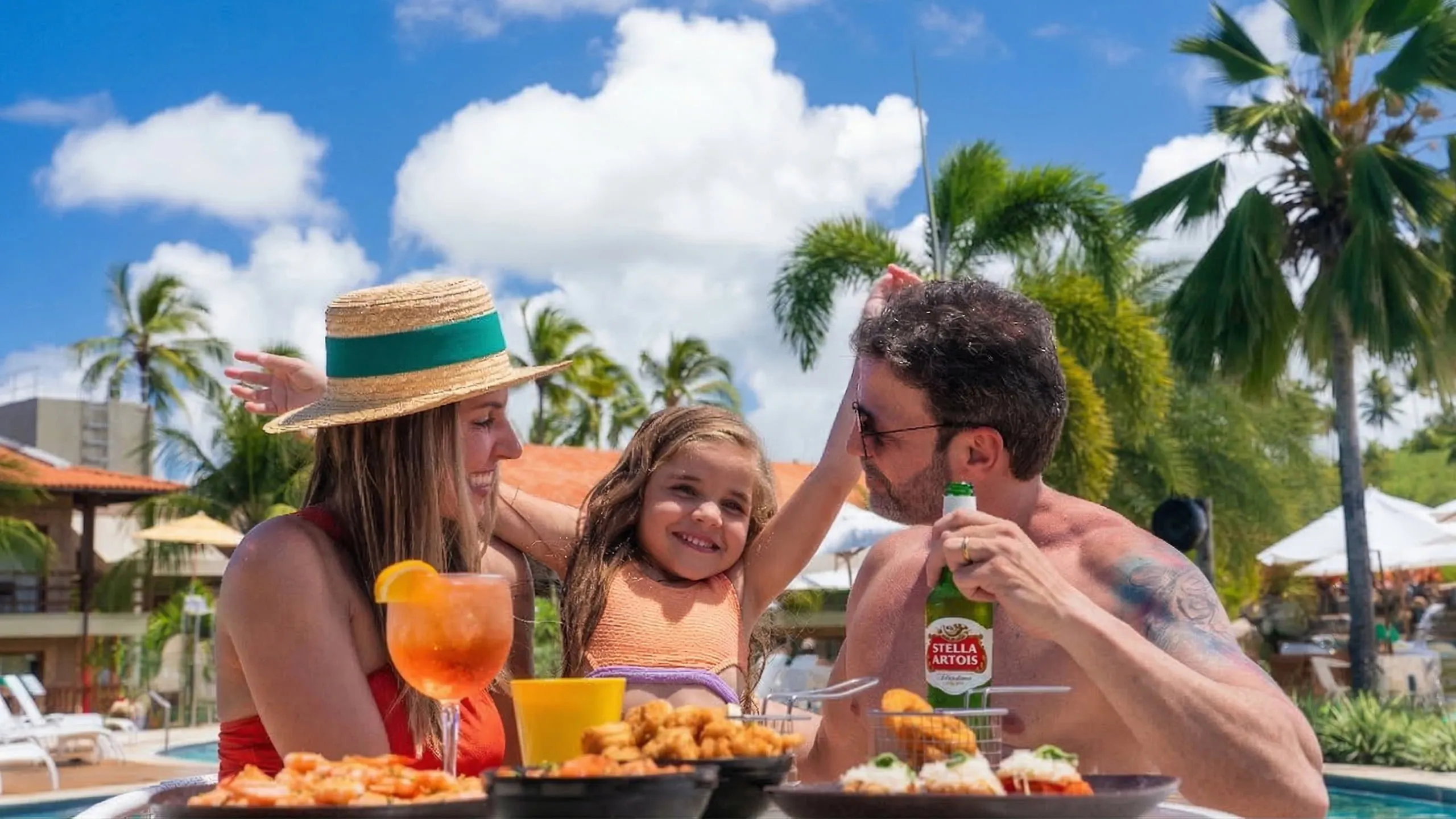 Família aproveitando o All Inclusive com petiscos, drinks e cerveja Stella Artois em área de lazer do Salinas Maragogi. E ao fundo coqueiros, espreguiçadeiras e dia ensolarado.