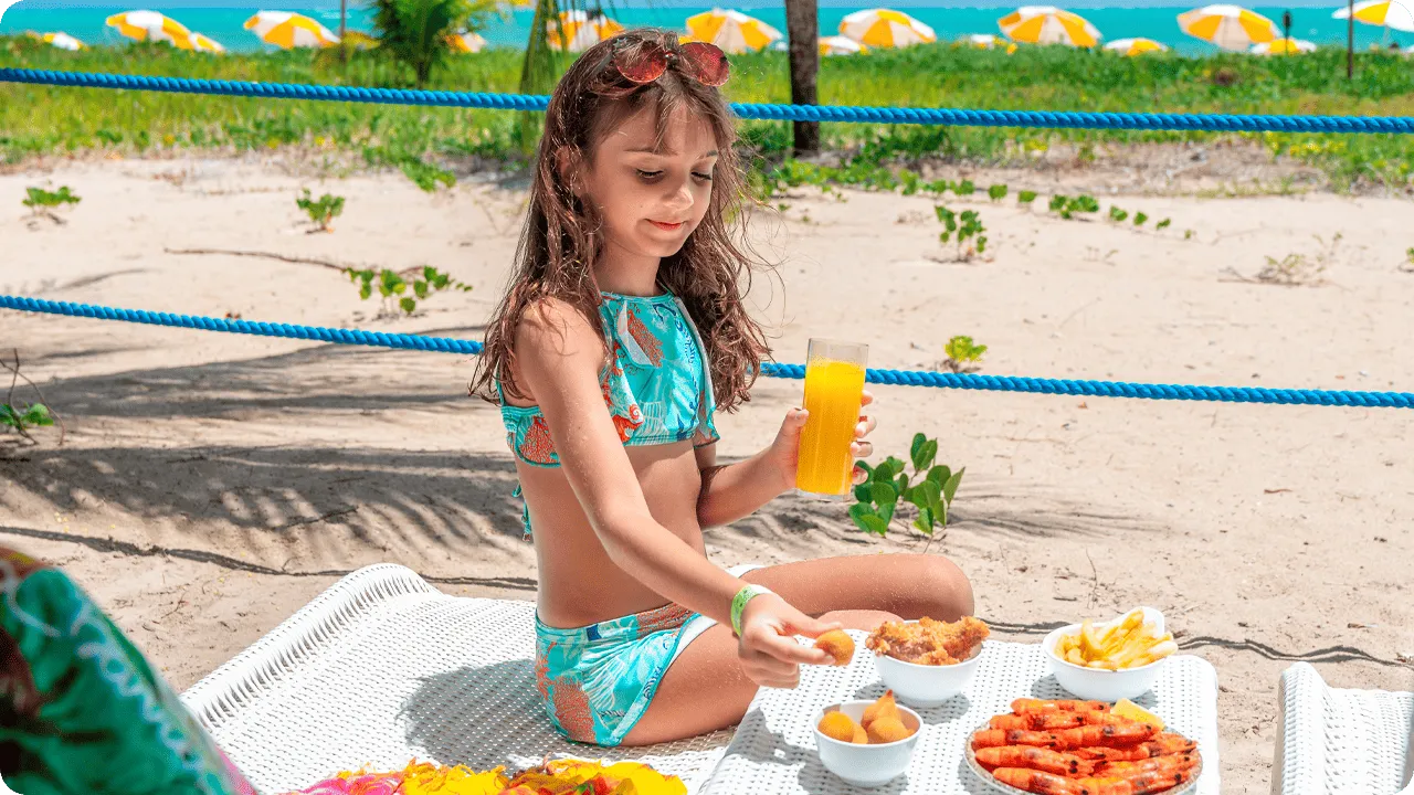 Criança aproveitando petiscos e suco na beira da praia em resort all inclusive.