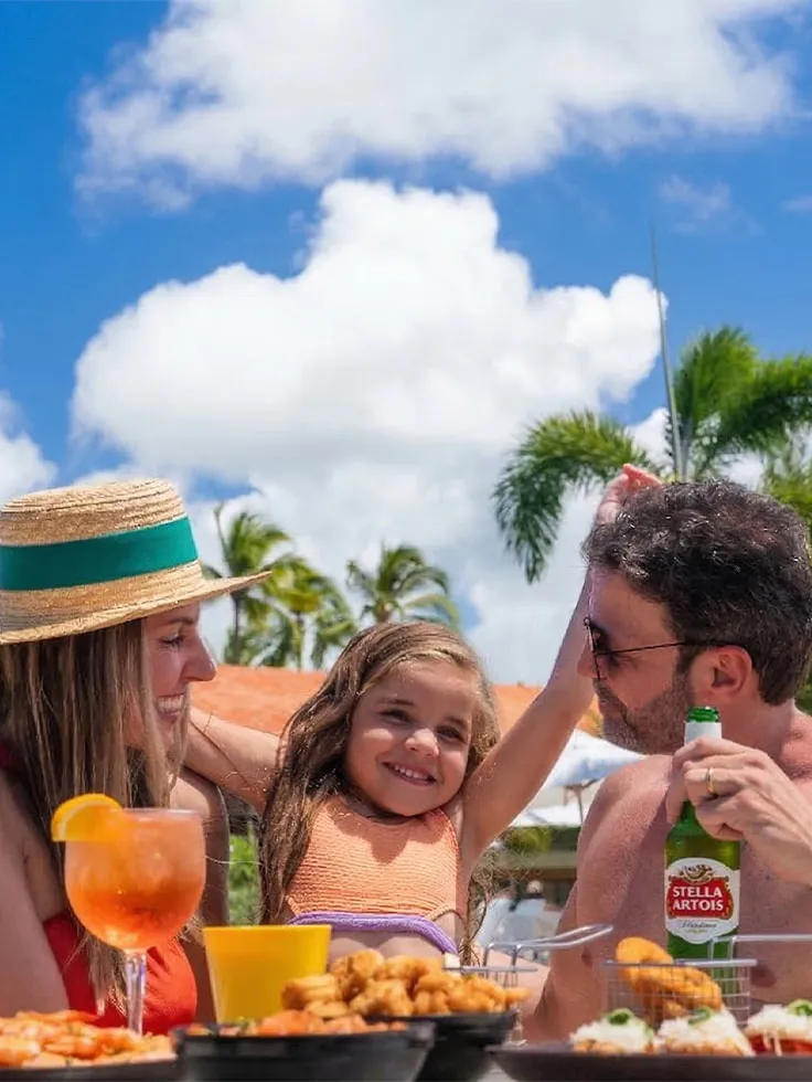 Família aproveitando o All Inclusive com petiscos, drinks e cerveja Stella Artois em área de lazer do Salinas Maragogi. E ao fundo coqueiros, espreguiçadeiras e dia ensolarado.