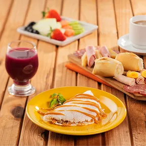 Mesa de café da manhã no resort, com tapioca doce, pães, frios, frutas frescas, suco e café, servidos em uma mesa de madeira.