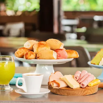 Mesa de café da manhã regional no Salinas Maceió, com destaque para um prato de cuscuz com carne de sol, acompanhado por suco, bolo, macaxeira, café e frutas.