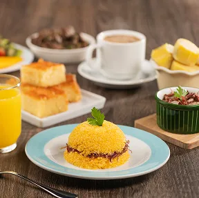 Mesa de café da manhã com um prato da sobremesa regional cartola, uma tigela de pães de queijo e uma fatia de bolo de rolo, com frutas e café ao fundo.