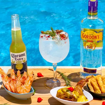 Bandeja na beira da piscina com uma variedade de petiscos, como camarão e isca de peixe, e bebidas, incluindo um drink de gin e uma cerveja Corona, com a água azul da piscina ao fundo.