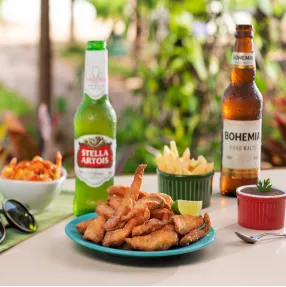 Mesa com um prato de isca de peixe frita, batata frita e molhos, ao lado de duas garrafas de cerveja, Stella Artois e Bohemia. A cena se passa em uma área externa com plantas ao fundo.