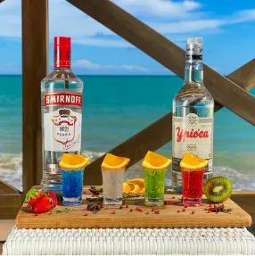 Bandeja de madeira com uma garrafa de vodka Smirnoff e uma de cachaça Ypióca ao lado de quatro shots coloridos enfeitados com laranja, morango e kiwi, tendo o mar de Maceió como cenário de fundo.