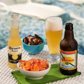 cervejas Corona Extra e Original em long neck ao lado de um copo com cerveja e também petiscos sendo camarão e carnes em fatia sobre mesa de área externa. Ao fundo espreguiçadeiras em parte rasa da piscina.