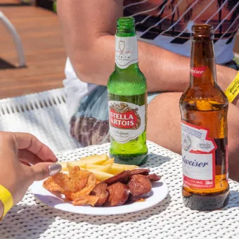 Bandeja de madeira na beira da piscina com três cervejas, Beck's, Brahma Duplo Malte e Stella Artois, e porções de petiscos, incluindo camarão frito e costelinha barbecue, com a água azul da piscina ao fundo.