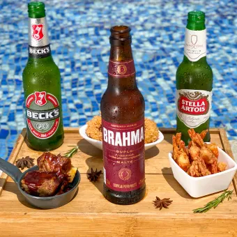Bandeja de madeira na beira da piscina com três cervejas, Beck's, Brahma Duplo Malte e Stella Artois, e porções de petiscos, incluindo camarão frito e costelinha barbecue, com a água azul da piscina ao fundo.