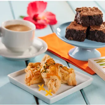 Mesa azul com uma xícara de café, um prato de folhados salgados e um suporte com brownies de chocolate. Uma mão segura a xícara e uma flor de hibisco decora o ambiente, representando uma pausa para o café no resort.