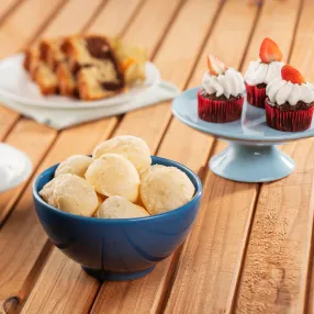 Mesa posta em uma superfície de madeira, com uma tigela azul de pães de queijo em destaque, além de cupcakes com morango, fatias de bolo mesclado e uma xícara de café preto ao fundo.