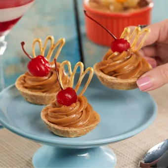 Mesa com diversas sobremesas finas, incluindo mini tortas de doce de leite, uma taça de mousse de chocolate e uma panna cotta de frutas vermelhas, com uma mão segurando uma das tortas.