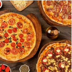 Vista de cima de uma mesa de madeira com várias pizzas, incluindo margherita, calabresa e uma pizza doce de chocolate com morango, cercadas por ingredientes frescos e um cortador de pizza.