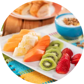 Café da manhã tropical com frutas frescas, suco verde, iogurte com granola e pães sobre toalha colorida.