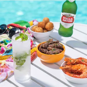 Mesa branca à beira da piscina com mojito no copo alto, cumbuca de carne em tiras, bolinhos fritos, camarões e uma mão segurando uma cerveja Stella Artois; óculos e canga colorida ao lado.