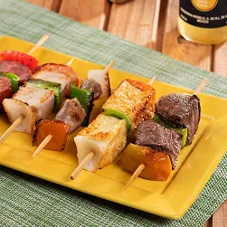 Prato amarelo com espetinhos variados de carne, queijo, linguiça e legumes sobre jogo americano verde, ao lado de panelinha com arroz, tigela de farofa e garrafa de cerveja Corona bem gelada sobre mesa de madeira.