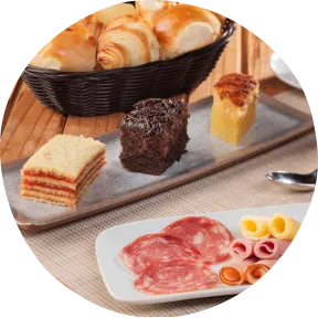Café da manhã completo com cesta de pães, frios, bolos variados, salada de frutas e café preto sobre a mesa.