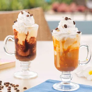 Duas taças de vidro com cafés gelados cobertos com chantilly e grãos de café, uma com calda de chocolate e outra com caramelo; ao fundo, ambiente de resort desfocado. Grãos de café espalhados e guardanapo azul sobre a mesa.