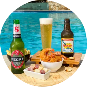 Petiscos variados com camarões empanados, batatas fritas, queijo com linguiça e cerveja gelada sobre bandeja de madeira à beira da piscina.