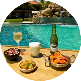 Petiscos variados com camarões empanados, batatas fritas, queijo com linguiça e cerveja gelada sobre bandeja de madeira à beira da piscina.