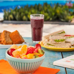 Tigela de salada de frutas, copo de suco roxo, sanduíches e fatias de bolo sobre mesa à beira da piscina.