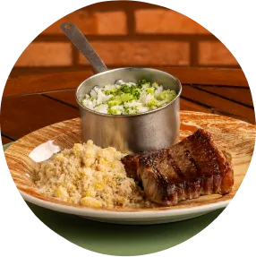 Prato com arroz, carne grelhada e salada fresca sobre mesa de madeira com fundo de tijolos e talheres ao lado.