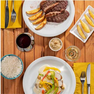 Bife grelhado com batatas e peixe ao molho com pimentões, arroz, farofa e taças de vinho sobre mesa de madeira.