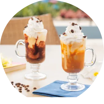 Cafés gelados com chantilly, calda de chocolate e caramelo servidos em canecas de vidro com grãos de café e flores decorativas.