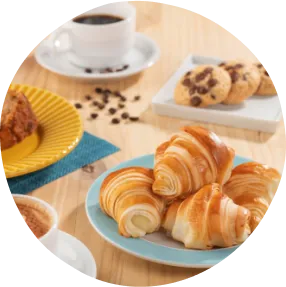 Uma mesa com cappuccino, croissants, bolo, cookies com pratos azul, branco e amarelo. Acompanhado de um café preto e flores decorativas.