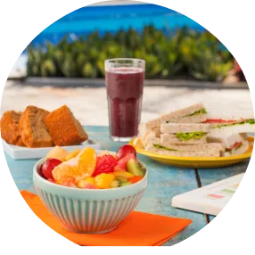 Café da tarde ao ar livre com frutas tropicais, sanduíches, bolo e smoothie roxo sobre mesa azul à beira da piscina.