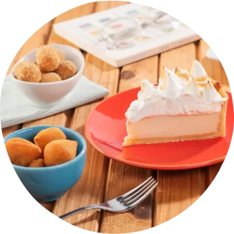 Uma torta, suco de laranja, coxinhas, bolinhos e puff pastry sobre mesa rústica com livro decorativo ao fundo.