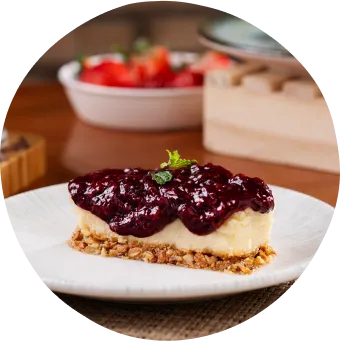 Sobremesa com cheesecake de frutas vermelhas em um prato, chocolate com raspas, morangos frescos e toque de hortelã em cima de uma mesa.