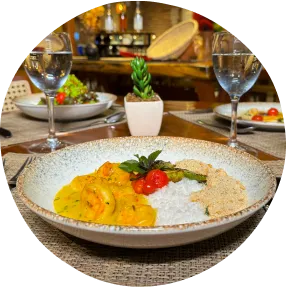 Jantar com camarões ao molho, arroz branco, farofa e taças de vinho branco servido em ambiente acolhedor com mesa posta pra três pessoas.