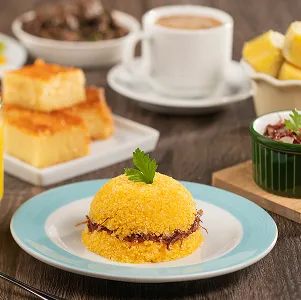Mesa com cuscuz recheado de carne seca, suco de laranja, café, bolo de milho, frutas e acompanhamentos típicos do nordeste.