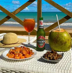 Mesa à beira da praia com camarões, petiscos, cerveja Beck's, coco gelado e drink colorido com vista para o mar