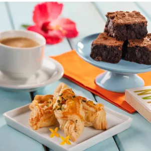Mesa com xícara de café, brownies de chocolate, folhados salgados e um livro ao fundo
