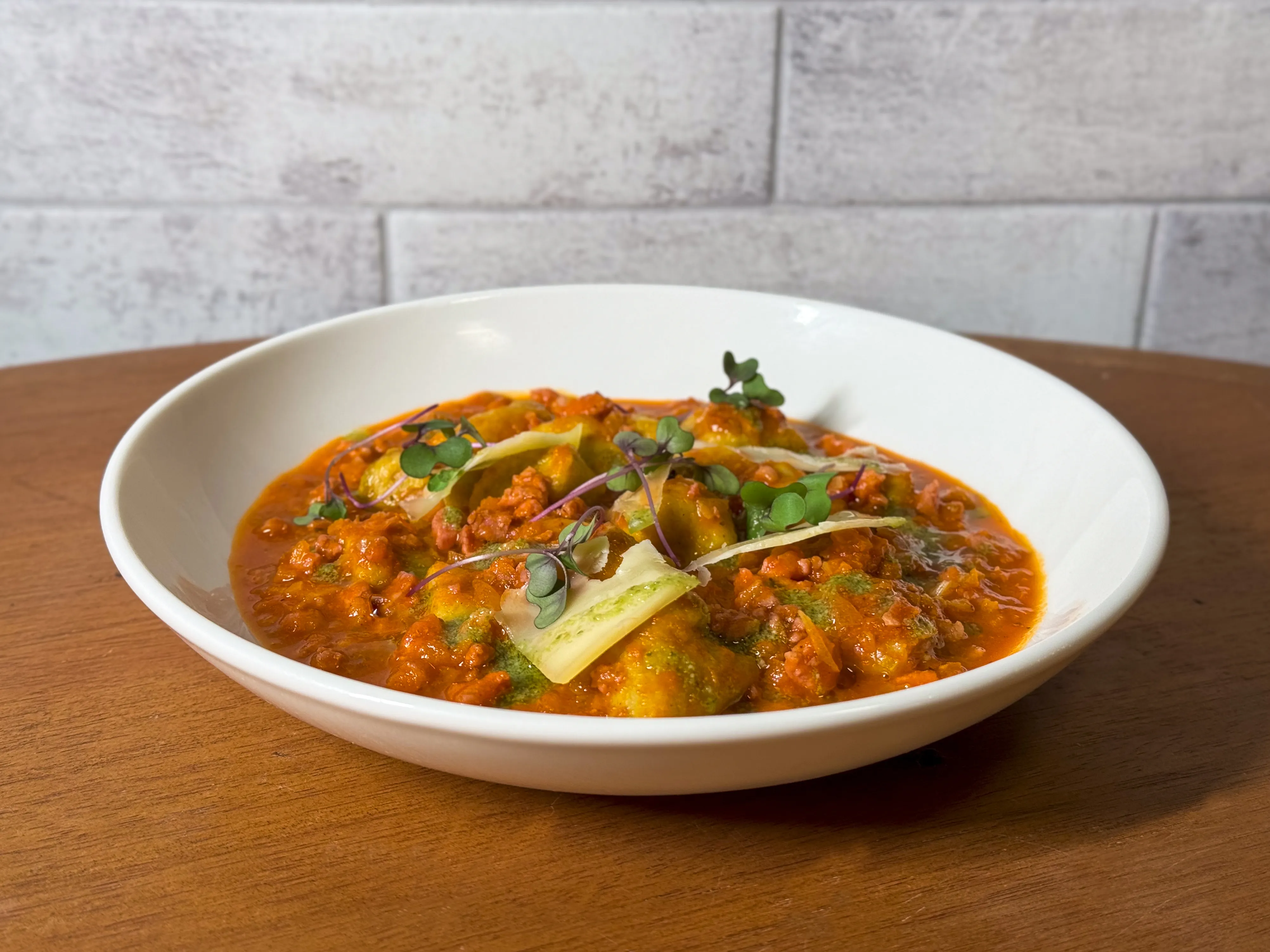 Prato de nhoque artesanal com molho bolonhesa encorpado finalizado com lascas de queijo parmesão e brotos de manjericão servido em bowl branco sobre mesa de madeira.