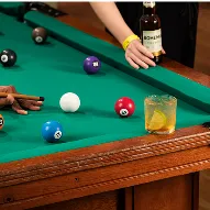 Mesa de sinuca com bolas coloridas, copo com drink de limão e pessoa segurando cerveja Bohemia