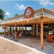 Bar de praia com cobertura de palha, mesas e cadeiras na areia, quiosques e vista direta para o mar.