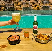 All Inclusive completo no Salinas Maragogi Bandeja à beira da piscina com Stella Artois, acompanhadas de petiscos como camarão e chips de batata doce, com cascata de pedras ao fundo.