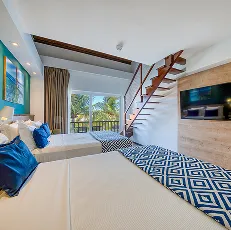 Vista do quarto Apartamento Suíte Familiar no Salinas Maceió, equipado com duas camas de casal, decoração moderna com parede azul, TV, ar-condicionado e uma escada pro mezanino. E ao fundo a varanda com vista pra área de lazer do resort.