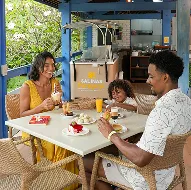 Família no Canoas Café aproveitando de lanches e cafés disponíveis, com bolos, doces, sucos. Ao fundo o espaço do do Canoas, e teto rústico com pilastras azuis.
