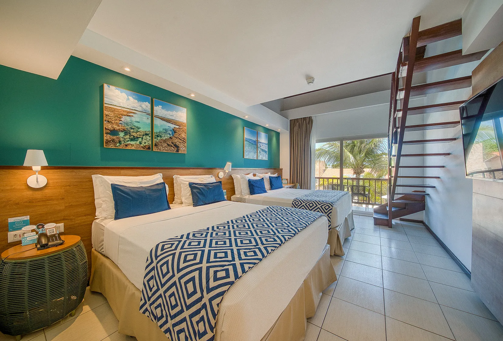 Suíte Familiar, uma das acomodações do Salinas Maceió All Inclusive Resort, com duas camas queen size aconchegantes, painel de madeira atrás delas, com luminária, porta da varanda e escada para o mezanino.